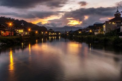 Illuminated buildings by lake against sky at sunset