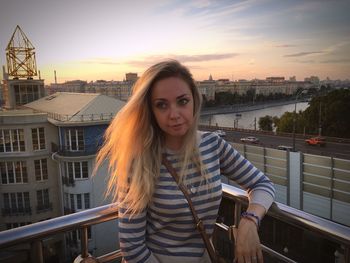 Portrait of beautiful woman standing on railing in city against sky during sunset