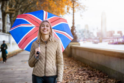 Portrait of smiling young woman holding umbrella while standing on footpath