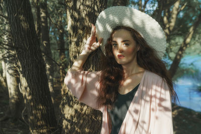 Portrait of young woman with hat standing against tree