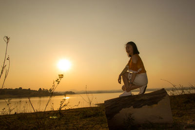 Woman crouching on log at lakeshore against clear sky during sunset
