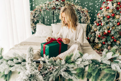 Lovely blonde young woman in pajamas smiling and untying ribbon on christmas gift 