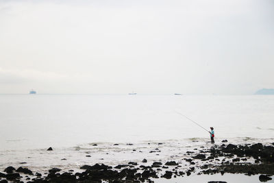 Rear view of man fishing in sea against clear sky