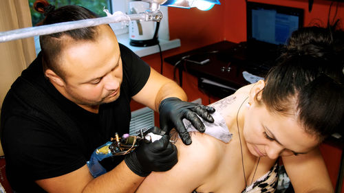 In a tattoo salon , a specialist is doing a tattoo on woman's back, a floral ornament. a man works