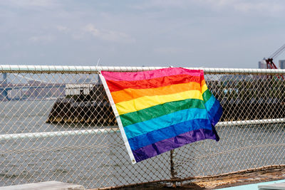 Rainbow flag hanging on chainlink fence against sky