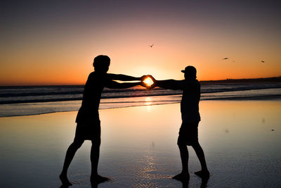 Silhouette of two men holding the sun on the beach at sunset