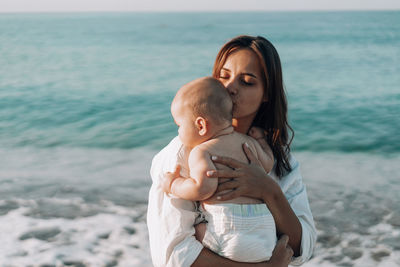 A young mother walks along the beach with a small child in diapers.