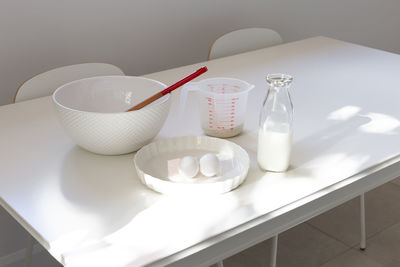 White cooking utensils and bowl, milk, eggs and flour in measuring cup set on table in dappled light