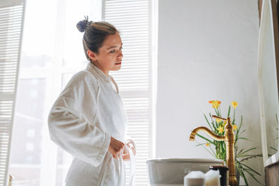 Young woman with dark long hair in white bathrobe near mirror in bathroom at home