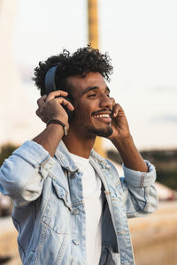 Close-up of smiling young man listening music through headphones against clear sky