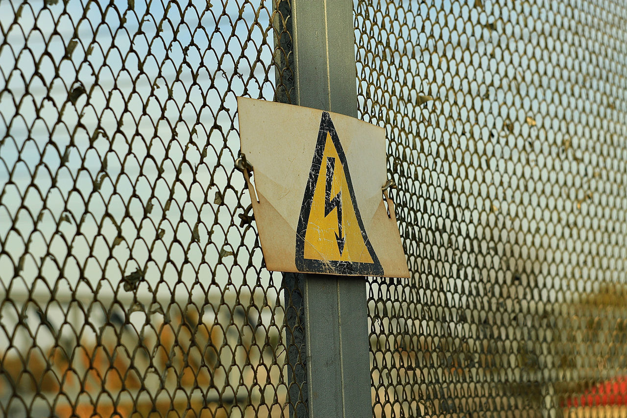 CLOSE-UP OF YELLOW SIGN ON METAL FENCE AGAINST BLURRED BACKGROUND