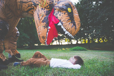 Boy lying on grassy land while playing with person wearing dinosaur costume in park