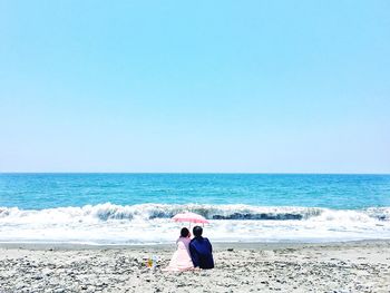 Rear view of couple with umbrella sitting on beach against clear sky