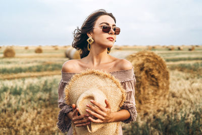 Smiling woman in sunglasses with bare shoulders on a background of wheat field and bales of hay.