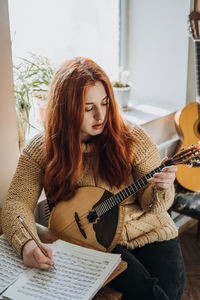 Redhead woman playing acoustic domra folk musical instrument and looking into note. young woman