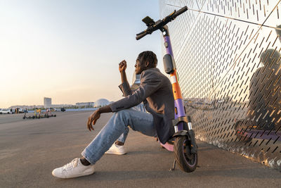 Smiling young businessman sitting on electric push scooter by metal wall