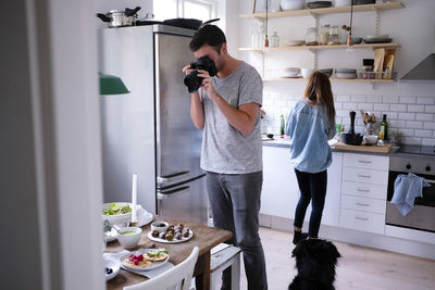 Male blogger photographing food through dslr against woman standing in kitchen