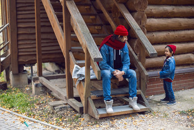 Man and his son, a boy in red hats, are sitting next to a wooden house in autumn