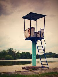 An old white and rusty metal lifeguard tower with chair on a pond beach.