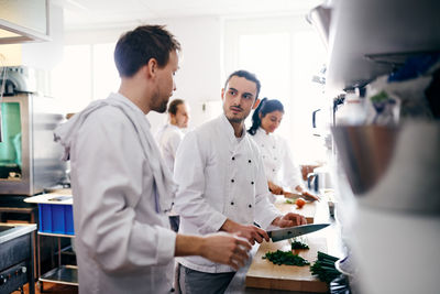 Young chef talking with colleague while chopping vegetable in commercial kitchen