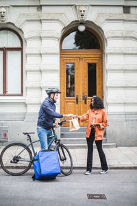 Food delivery man delivering package to woman standing on sidewalk in city