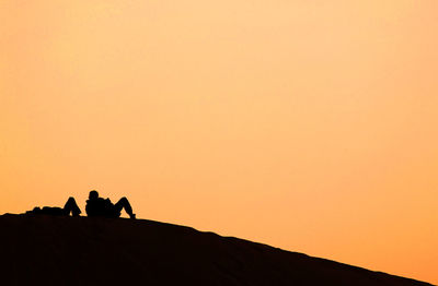 Silhouette people resting on sand dune at erg chebbi desert against sky during sunset