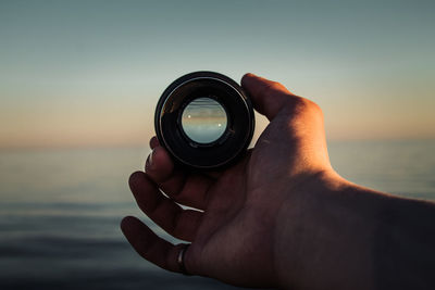 Close-up of hand holding camera against sky during sunset