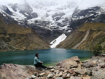 Side view of woman sitting by lake and mountains during winter