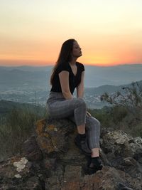 Young woman sitting on rock against sky during sunset