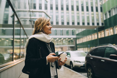 Businesswoman with coffee and smart phone looking away while standing on sidewalk against modern building in city