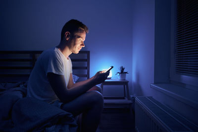 Lonely man sitting on bed in bedroom and using phone at night. 