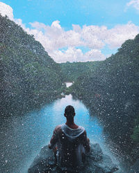 Rear view of man sitting on rock over river against sky