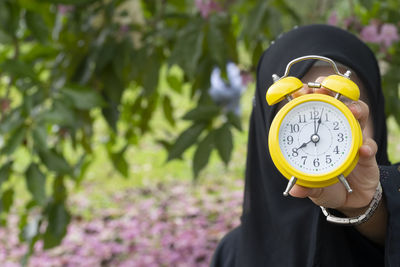 Close-up of person holding yellow alarm clock against trees