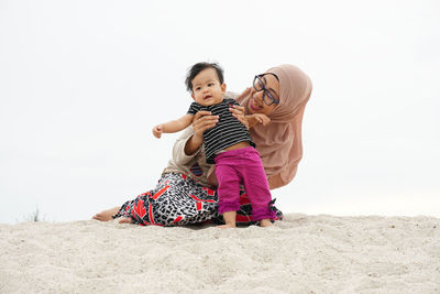 Mother wearing hijab holding cute daughter while sitting on sand at beach against sky