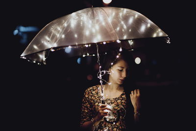 Woman looking down while holding illuminated umbrella while standing outdoors at night