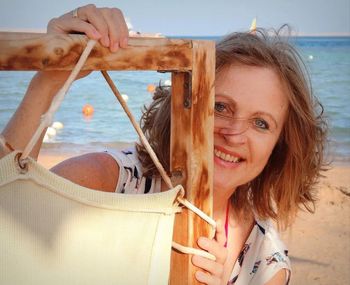 Portrait of happy mature woman standing behind wooden frame at beach