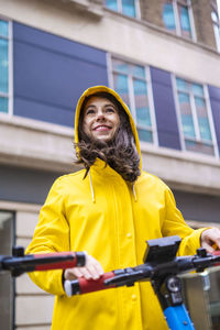 Happy young woman standing with electric push scooter in front of building