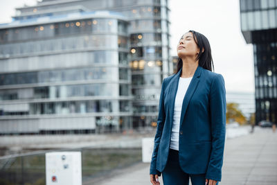 Businesswoman standing with eyes closed near buildings