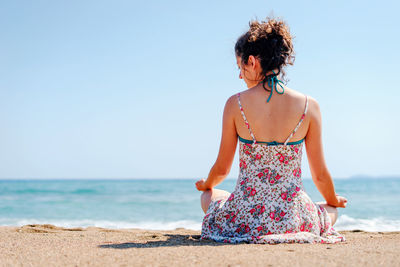 Rear view of woman meditating while sitting at beach against clear sky