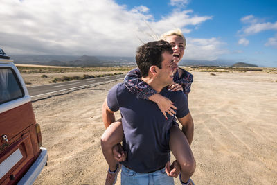 Happy man carrying woman while walking at beach against sky