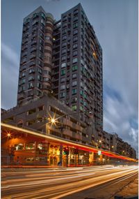 Light trails on road by buildings against sky at night