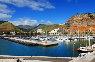 Sailboats moored at harbor by mountains against blue sky