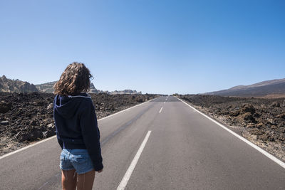 Rear view of young woman standing on road against clear blue sky during sunny day