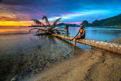 Man with tripod sitting on fallen tree at beach against sky