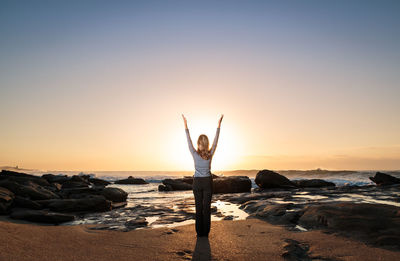 Rear view of woman exercising yoga with hands raised on shore at beach against sky during sunrise