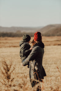 Child in knitted warm green hat inhis mother arms in maroon hat , warmly hugging  inautumn landscape 