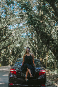 Portrait of young woman standing by car against trees