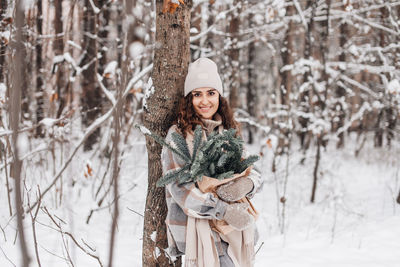 A stylish young woman in a hat stands in a snowy forest with branches of a nobilis fir near a tree