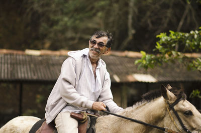 Smiling man in sunglasses riding horse against tree