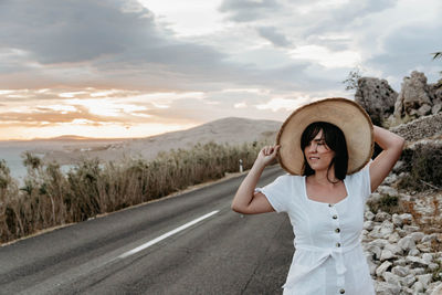 Portrait of beautiful young woman standing by scenic panoramic road. summer, sunset.